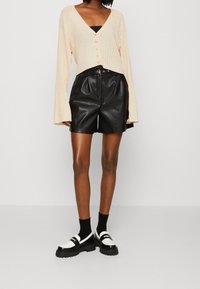 Cardigan beige côtelé court avec un décolleté en V et des détails de boutons, associé à un short noir en simili cuir taille haute et des chaussures noires et blanches épaisses.
