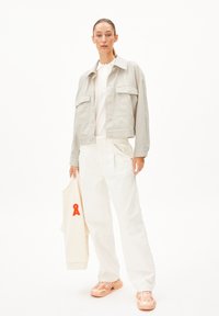 Frau trägt eine hellbeige Jacke, weiße Hose und blassrosa Sneakers und hält ein cremefarbenes Sweatshirt mit einem roten Grafik in ihrer linken Hand.