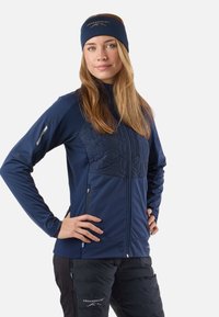 Giacca softshell blu navy con design trapuntato sul petto, tasche con cerniera e fascia per capelli abbinata. Tessuto strutturato con taglio aderente.