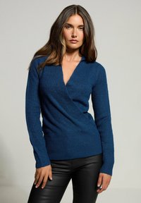 Donna che indossa un maglione V-neck blu scuro e strutturato e pantaloni in pelle nera, in piedi contro uno sfondo grigio chiaro uniforme.