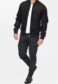 Veste bomber en suède noire avec col côtelé, associée à un pantalon cargo noir ajusté doté de poches latérales et de poignets élastiques. Des baskets noires terminent le look.