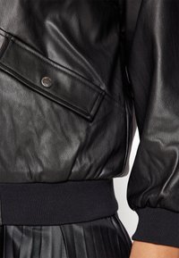 Svart läderjacka med en slät textur, med en sidoficka med en metallknäppning och ribbade svarta manschetter. Plisserad kjol synlig nedanför.