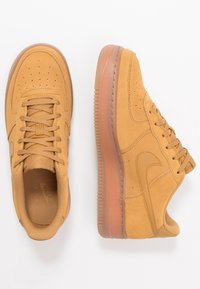 Zapatillas de ante marrón con puntera perforada, diseño con cordones y suela de goma. Presentan un detalle de costura visible y un logo en relieve.