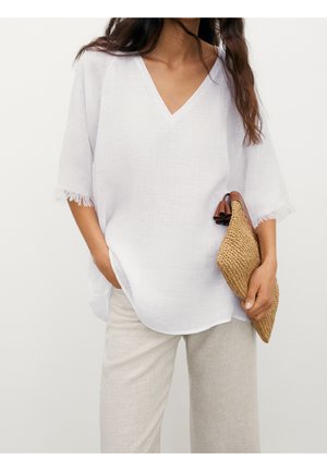 Woman wearing loose white V-neck top with fringed sleeves and light beige pants, holding a woven tan clutch bag.