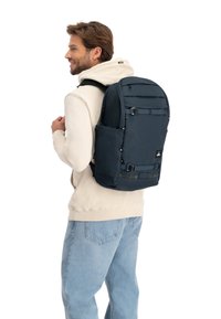 Maritimer Rucksack mit mehreren Fächern, verstellbaren Trägern und einem strukturierten Stoff, getragen über einem hellen Hoodie vor einem neutralen Hintergrund.