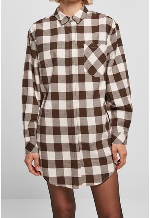 Vrouw draagt een bruin-wit geruite jurk met lange mouwen en een voorzak, gecombineerd met zwarte doorschijnende panty's, staand tegen een effen achtergrond.