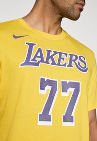 Κίτρινο βαμβακερό μπλουζάκι με τη λέξη «LAKERS» σε έντονα μωβ γράμματα κ αι τον αριθμό «77» με σχέδιο κάτω από αυτά. Λογότυπο Nike σε μωβ.