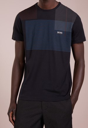 Homme portant un T-shirt noir avec des motifs rectangulaires multicolores en pointillés et le logo "BOSS" sur la poitrine, associé à un pantalon noir à pois.
