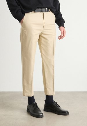 Mann trägt beige, gerade geschnittene Hose mit schwarzem Gürtel, schwarzen Socken und schwarzen Anzugschuhen, steht auf einem grauen Boden vor einer weißen Wand.