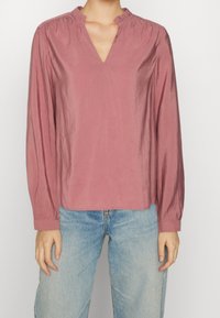Blusa rosa con scollatura a V e collo arricciato. Maniche lunghe e tessuto liscio, leggermente lucido. Abbinata a jeans azzurri chiari.