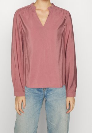 Blusa rosa con scollatura a V e collo arricciato. Maniche lunghe e tessuto liscio, leggermente lucido. Abbinata a jeans azzurri chiari.