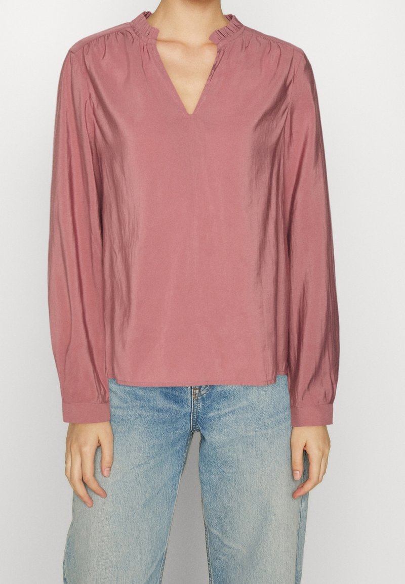 Blusa rosa con scollatura a V e collo arricciato. Maniche lunghe e tessuto liscio, leggermente lucido. Abbinata a jeans azzurri chiari.