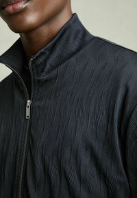 Veste noire zippée avec un motif ondulé texturé, col haut et détail de fermeture éclair métallique, mettant en avant son design épuré et son tissu.