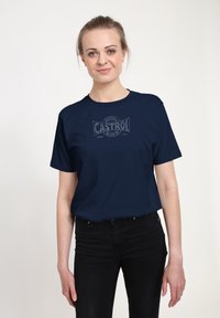 Námořnická modrá bavlněná tričko s šedým logem CASTROL, kulatým výstřihem a krátkými rukávy. Nosí se s černými džíny, což podtrhuje ležérní styl.
