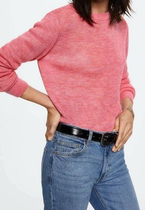 Personne portant un pull à manches longues rose et un jean bleu avec une ceinture noire, les mains posées sur les hanches, sur un fond clair uni.