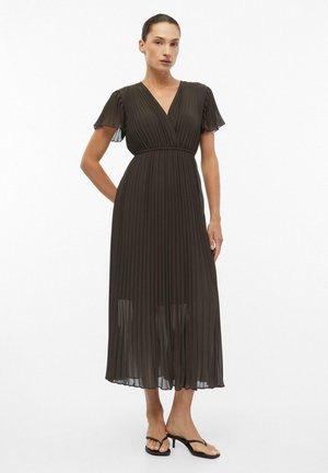 Femme portant une robe midi plissée noire avec des manches courtes volantées et un décolleté en V, associée à des sandales noires à brides et talons bas.