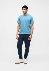 Άνδρας που φοράει ανοιχτό μπλε μπλουζάκι, σκούρο μπλε παντελόνι φόρμας, λευκά αθλητικά παπούτσια με μπεζ κάλτσες, στέκεται μπροστά σε απλό φόντο.