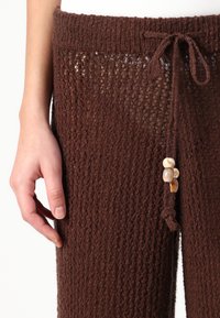 Pantalones de punto marrón con un patrón texturizado, que cuentan con una cintura ajustable con cordón y cuentas en forma de concha decorativas en el costado.