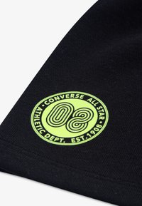 Μαύρο ύφασμα με στρογγυλό, νέον πράσινο λογότυπο. Το λογότυπο περιλαμβάνει τις λέξεις «CONVERSE ALL STAR», «ATHLETIC DEPT.» και τον αριθμό «08».