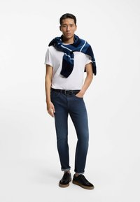 Valkoinen t-paita, tummansininen neule harteilla, tummansiniset farkut, mustat lenkkarit, joissa vaaleanruskea pohja, seisoo neutraalilla taustalla.