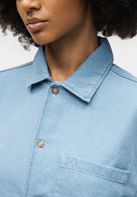 Κοντινό πλάνο ενός ατόμου που φοράει ανοιχτό μπλε πουκάμισο τζιν με γιακά, δύο εμφανή κουμπιά και μια τσέπη στο στήθος στη δεξιά πλευρά.