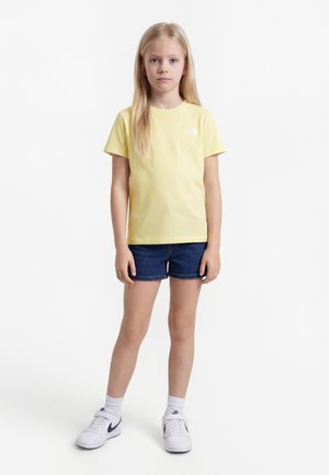 Ung pige med langt blondt hår iført en gul T-shirt, mørke denimshorts, hvide sokker og hvide sneakers, stående mod en hvid baggrund.