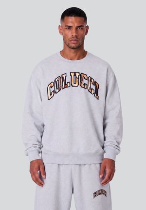 Uomo che indossa felpa grigio chiaro e pantaloni della tuta abbinati con testo "Colucci" in grandi lettere decorate sul petto e più piccole sui pantaloni.