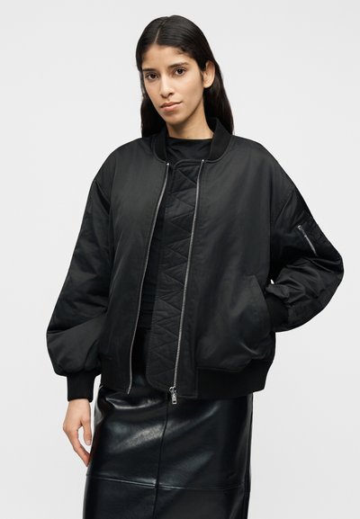 Fekete bombázó dzseki sima, fényes felülettel, bordázott mandzsettákkal és cipzáras záródással. Texturált fekete felső és bőrszoknya fölött viselve.