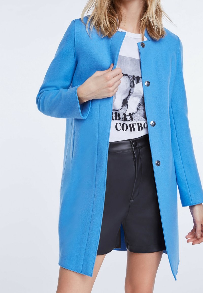 Femme portant un manteau boutonné bleu vif sur un t-shirt blanc graphique "Urban Cowboy" et un short en cuir noir, debout devant un fond uni.