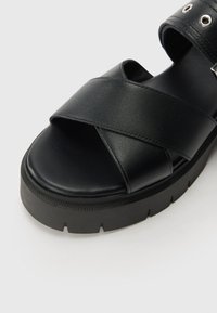 Black leather sandal with cross straps and a thick black rubber sole. Features perforated accents and a cushioned footbed for comfort.