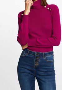 Femme portant un pull à col roulé moulant magenta avec des boutons aux épaules et un jean bleu taille haute avec cinq boutons visibles.
