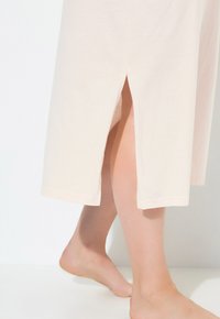 Abito in cotone color pesca pallido con spacco laterale, lunghezza al ginocchio, orlo dritto e maniche corte, su un piede nudo che poggia su una superficie bianca.