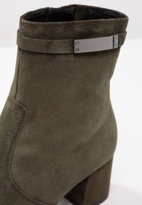 Botín de ante en verde oliva con tacón cuadrado. Presenta un acento de hardware plateado en el cuello y detalles de costura sutiles.