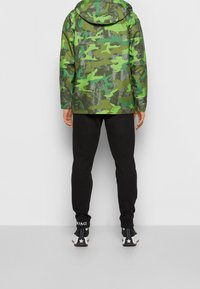 Camouflage jacket in green tones with a hood, worn over black pants. Features a textured design with a mix of dark and light green patterns.