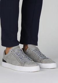 Zapatillas de ante gris con suelas de goma blanca, que presentan cordones planos y un acento negro en el talón. Detalles de costura visibles.