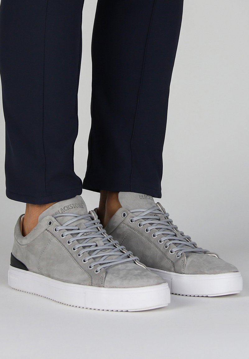Zapatillas de ante gris con suelas de goma blanca, que presentan cordones planos y un acento negro en el talón. Detalles de costura visibles.