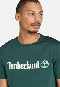 Zelené bavlněné tričko s bílým logem "Timberland" a emblémem stromu, krátké rukávy a design s kulatým výstřihem.