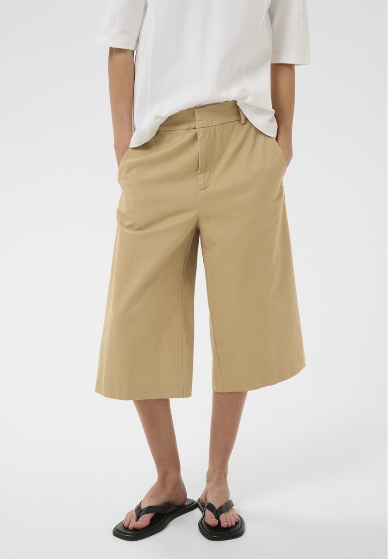 Personne portant un short beige large arrivant aux genoux, une chemise blanche à manches courtes et des sandales noires, debout les mains dans les poches.