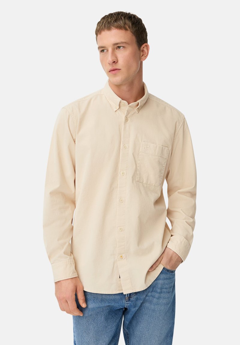 Beige Hemd aus Baumwolle mit Knopfleiste, Brusttasche, klassischem Kragen und langen Ärmeln. Getragen mit blauen Jeans.