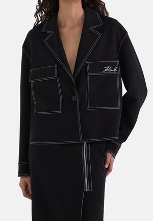 Blazer noir avec coutures contrastantes blanches, deux poches avant à rabat, dont une brodée "Karl", porté avec une jupe assortie dotée d'une fermeture éclair devant.