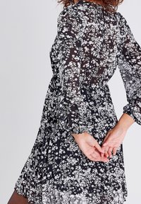 Robe noire à fleurs avec tissu transparent, manches longues et taille froncée. Présente une silhouette fluide avec des motifs de fleurs blanches sur toute la surface.