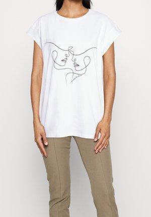 Personne portant un t-shirt blanc avec un dessin en ligne continue noire de deux visages et un texte indiquant "proceed to love", associé à un pantalon beige.