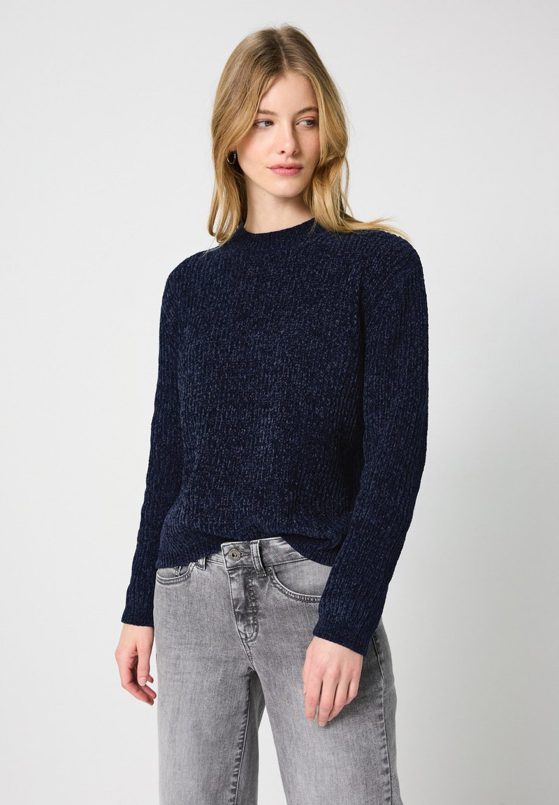 Marineblauer, strukturierter Strickpullover mit langen Ärmeln und hohem Kragen, kombiniert mit hellgrauen Denim-Jeans, die einen lässigen Schnitt zeigen.