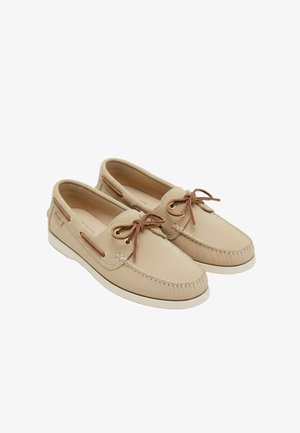 Scarpe mocassino da barca in pelle beige con lacci in pelle marrone, suole bianche e cuciture stile mocassino su uno sfondo bianco.