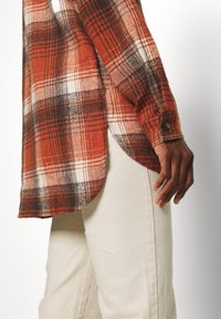 Rood en oranje geruit flanellen overhemd met een afgeronde zoom en mouwen met knopen, gecombineerd met lichtgekleurde, gestructureerde broek.