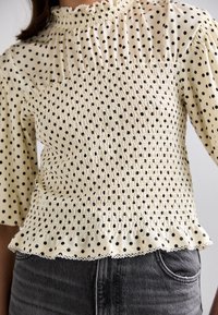Femme portant un chemisier à manches longues couleur crème à pois noirs, taille smockée, ourlet à volants, associé à un jean gris foncé.