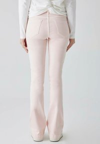 Jeans de mezclilla de corte acampanado en rosa claro, con una cintura de altura media, dos bolsillos traseros y una textura deslavada. Combinados con una blusa blanca de manga larga.