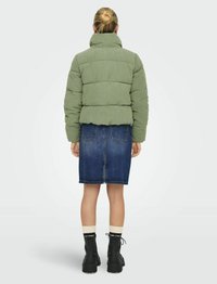 Veste matelassée en velours côtelé vert, au design court. Portée sur une jupe en denim, associée à des bottines noires et des chaussettes crème. Vue de dos.