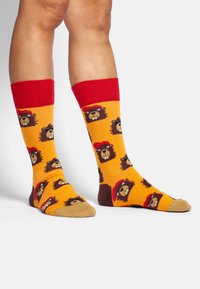 Calcetines amarillos con puños rojos que presentan caras de león de dibujos animados repetidas, con puntas marrones y un material suave y elástico.