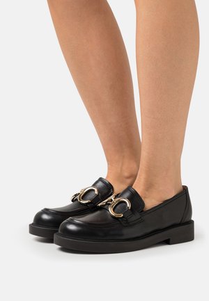 Black leather loafers with a rounded toe, featuring gold hardware accents and a chunky black sole. Smooth texture with a minimalist design.